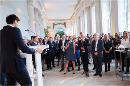 A speaker addresses a well-dressed crowd in an elegant room with large windows.