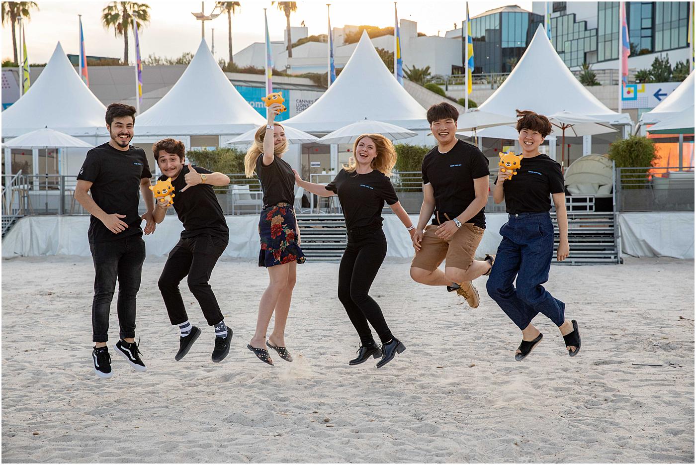 A group of people jumps excitedly on a sandy beach holding plush toys, with a row of white tents in the background.