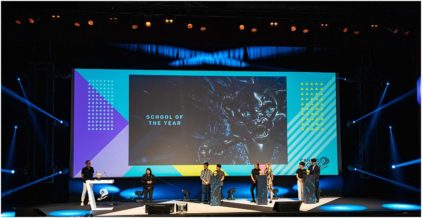 A group of people stands on stage at an event with a large screen displaying 