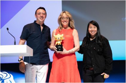 Three people smiling on stage, with one holding a yellow award statue.