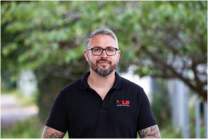 Man with tattoos and glasses wearing a black polo shirt stands outdoors.