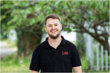 A person wearing a black polo shirt stands smiling outdoors.