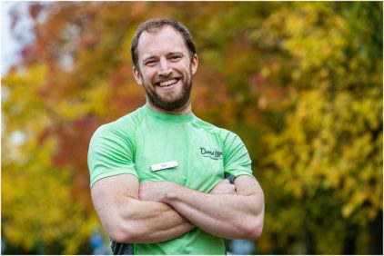 A man in a green shirt stands smiling with arms crossed against a background of colorful autumn leaves.