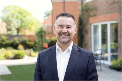 A man in a suit smiles outdoors in a garden setting.