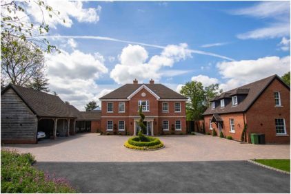 A large, brick house with adjacent smaller buildings and a circular driveway under a blue sky.