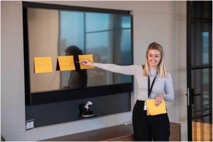 A woman smiles while pointing at sticky notes on a screen.