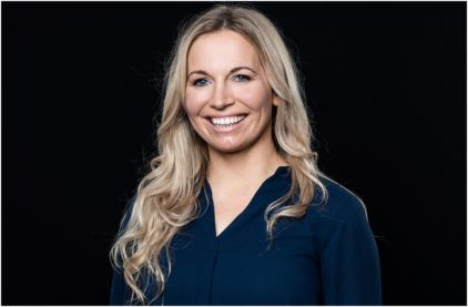 A smiling woman with long blonde hair wearing a dark blue top against a black background.