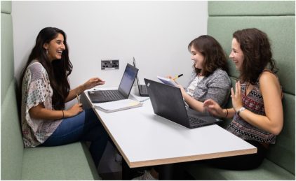 Three people are smiling and talking while sitting at a table with laptops.