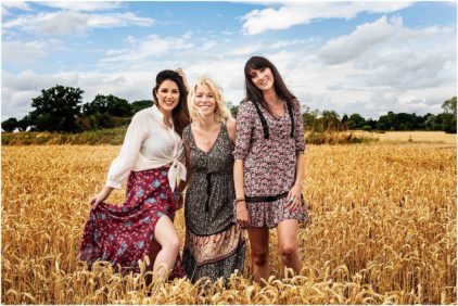 Three women smiling in a wheat field under a partly cloudy sky.