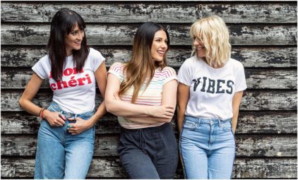 Three women smiling and standing against a rustic wooden wall.