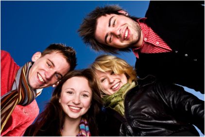 Four friends smiling and leaning in together against a clear blue sky.