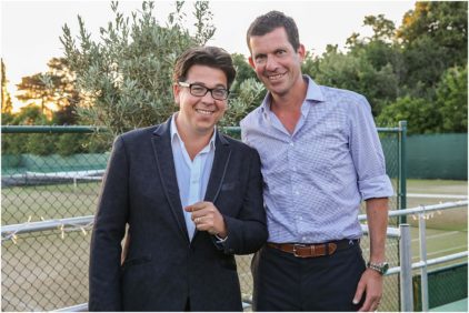 Two men are smiling and standing together outdoors in front of a tennis court.