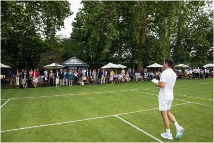 Outdoors image of a person holding a microphone on a tennis court with a crowd in the background.