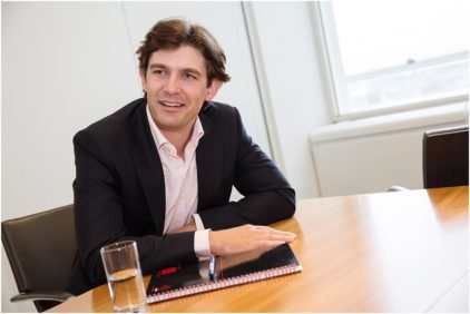 A man in a suit sits at a table with a notebook and glass of water, in an indoor office setting.