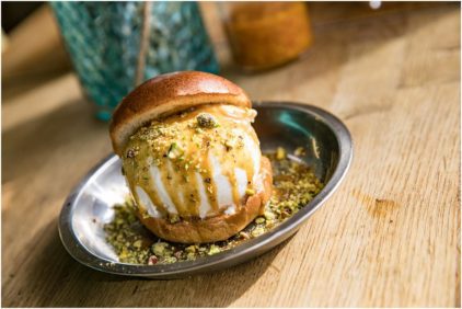 A close-up food image of an ice cream sandwich topped with pistachios on a metal plate.