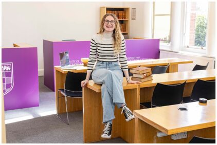 Portrait of a person sitting on a desk in a classroom setting with books and a laptop nearby.