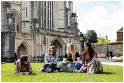 A group of four young adults sitting outdoors on grass, reading and chatting near a historic building.
