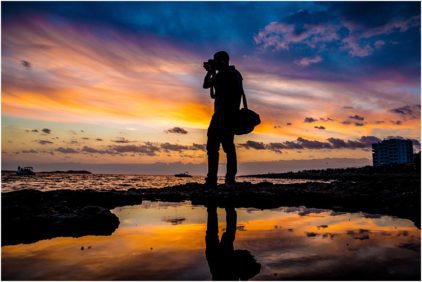 Silhouette of a person taking photos by the sea at sunset, with colorful skies reflected in the water, outdoors.