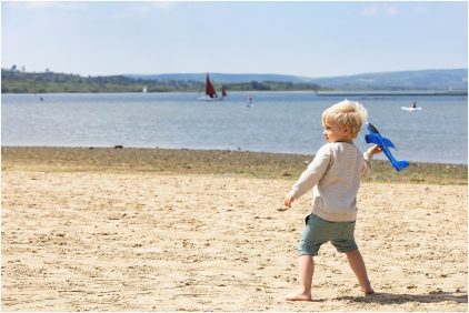 A young child plays with a toy airplane on a sandy beach, with a lake and sailboats in the background.