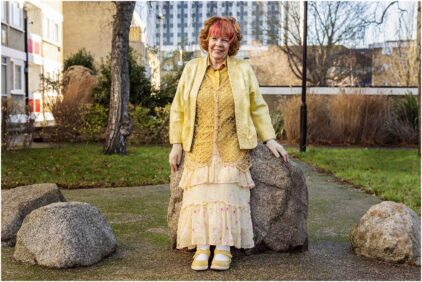 An elderly woman with colorful attire smiles while standing outdoors next to large rocks.