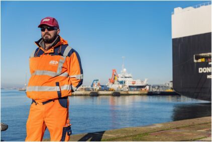 Outdoor portrait of a man in an orange safety uniform and cap standing near a harbor.