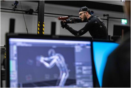 An indoor image of a person in motion capture gear aiming a mock weapon, with computer screens in the foreground.