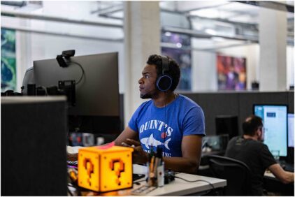 Indoors image of a person wearing headphones and working at a computer with a lit question mark cube nearby.