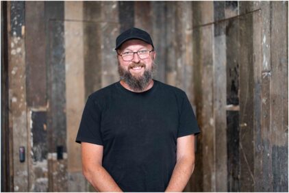 A smiling man in a black t-shirt and cap stands indoors against a wooden background.