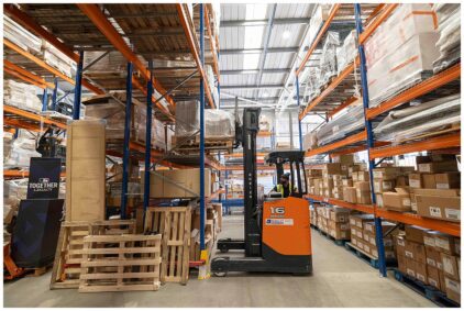 Interior of a warehouse with a worker operating a forklift among stacked shelves of boxes.