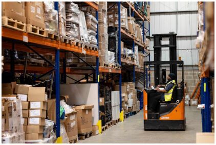 An indoor image of a worker operating a forklift in a warehouse aisle filled with stacked boxes.