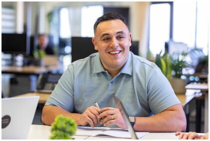 Smiling man in a light blue shirt sitting at a desk indoors.