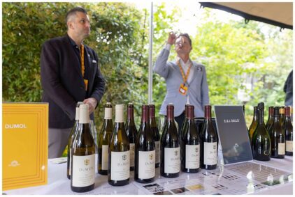 Outdoors image of a wine tasting event with bottles displayed on a table and two people standing behind.