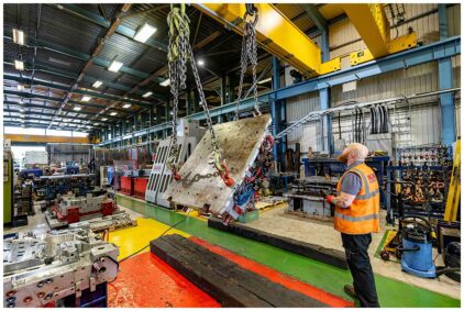 Indoors image of a worker observing a large metal piece being lifted by chains in a factory setting.