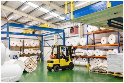 Indoors image of a warehouse with a forklift and large rolls of material on shelves.