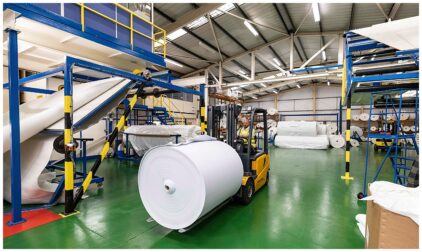 Indoor image of a warehouse with a worker operating a forklift carrying a large roll of material.